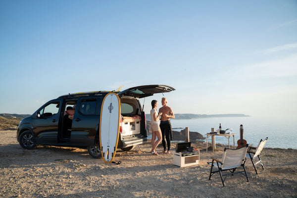 Paar liegt im Campingausbau eines Hochdachkombis am Strand mit Hund und Surfbrett