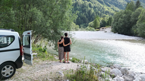 Paar am türkisfarbenen Soča-Fluss neben weißem Dacia Dokker vor grüner Bergkulisse