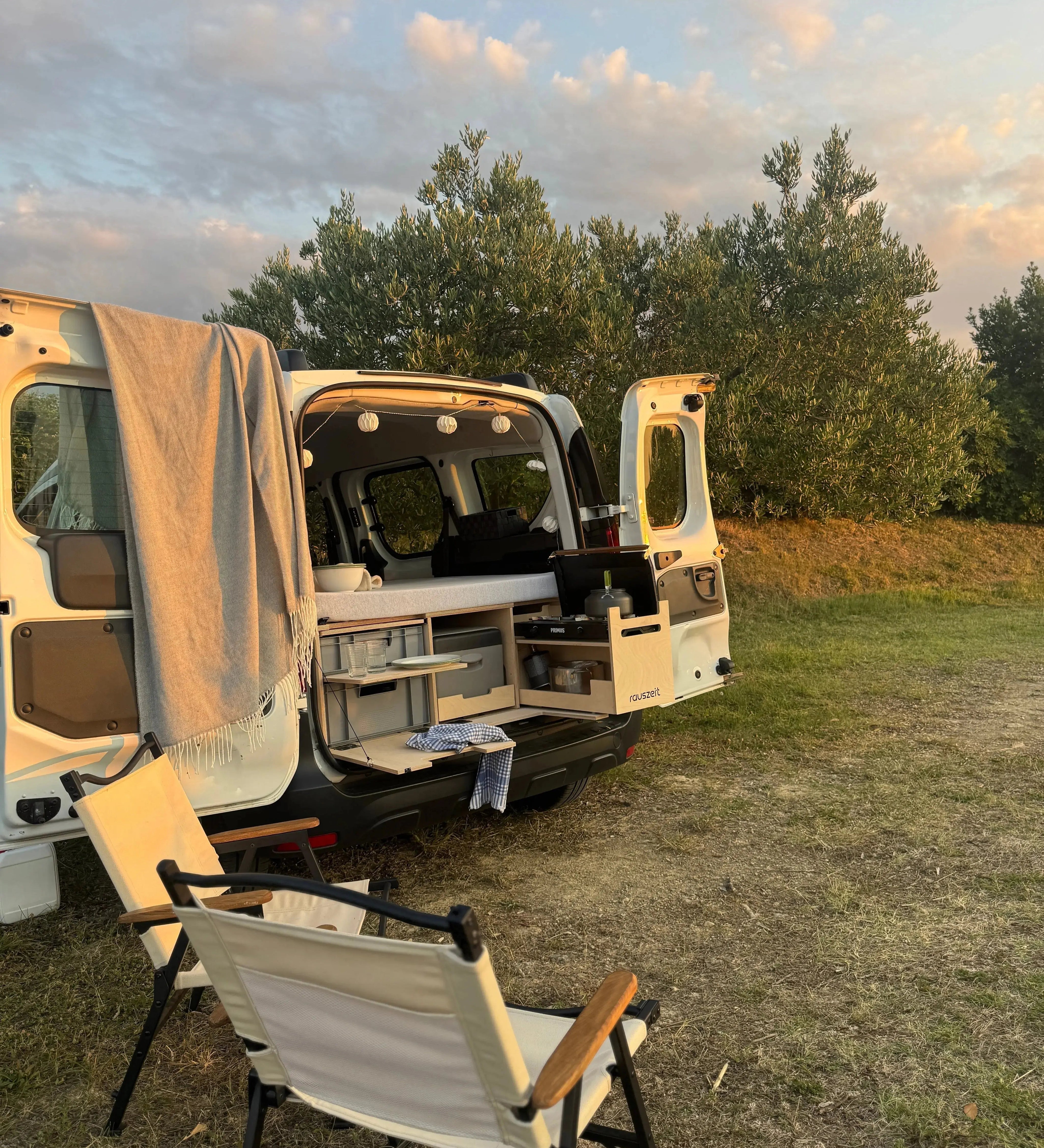 Campingbox im Hochdachkombi mit geöffneten Hecktüren und Campingstühlen bei Abendstimmung