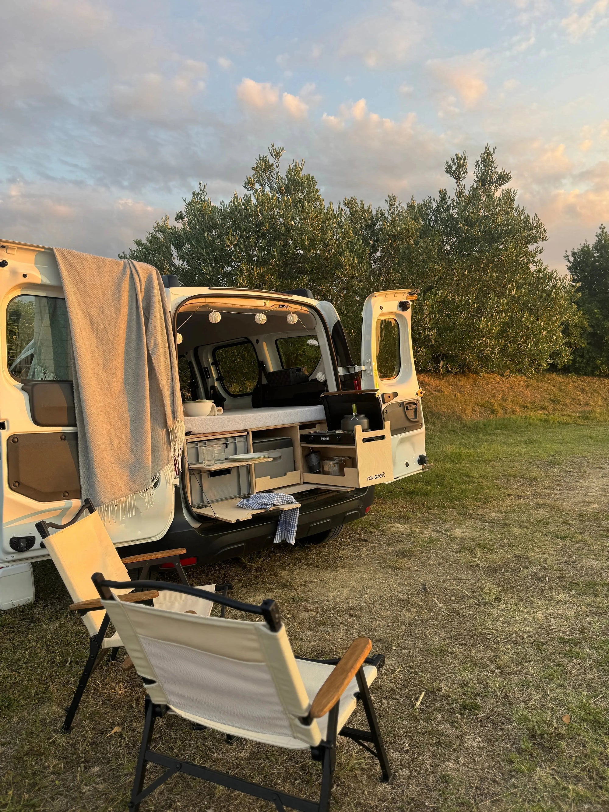 Campingbox im Hochdachkombi mit geöffneten Hecktüren und Campingstühlen bei Abendstimmung