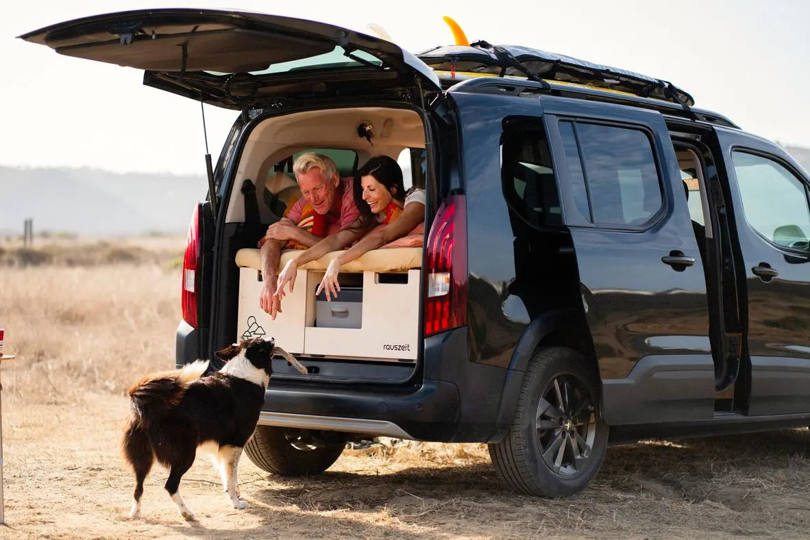 Paar liegt im Campingausbau eines Hochdachkombis am Strand mit Hund und Surfbrett