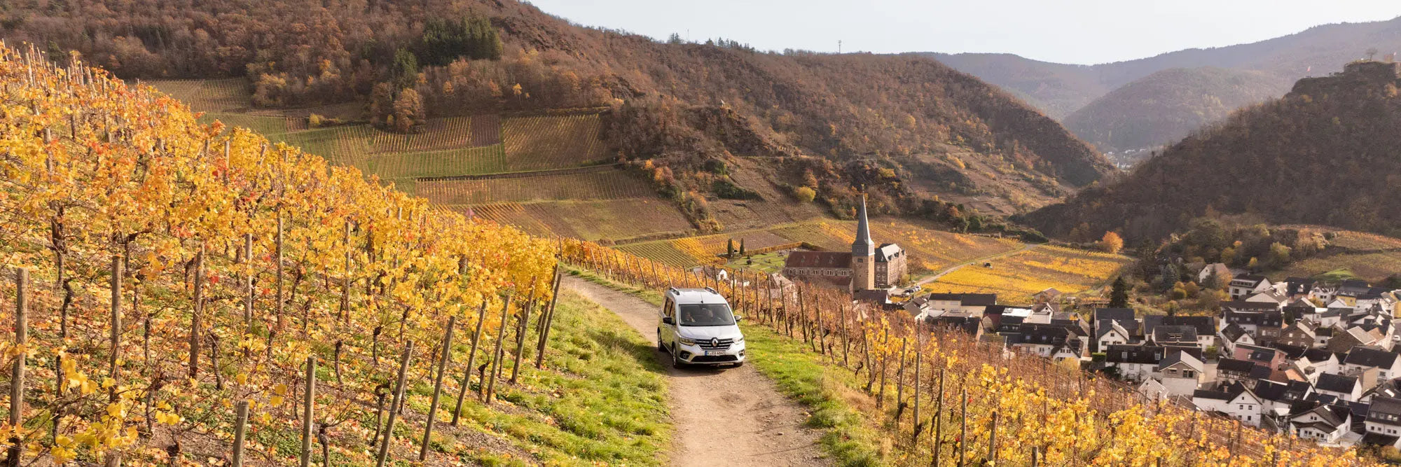 Hochdachkombi auf schmalem Weg zwischen herbstlichen Weinbergen im Flusstal