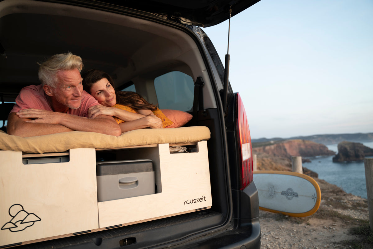 Paar liegt auf dem Schiebebett der Rauszeit Campingbox im Heck, darunter Kühlbox und Euroboxen, im Hintergrund die Küste