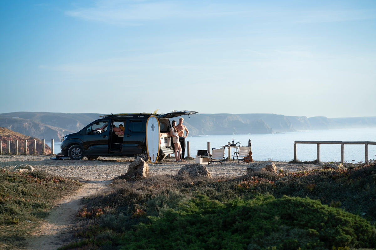 Hochdachkombi mit offener Heckklappe und Surfbrett an der Steilküste, daneben ein Paar