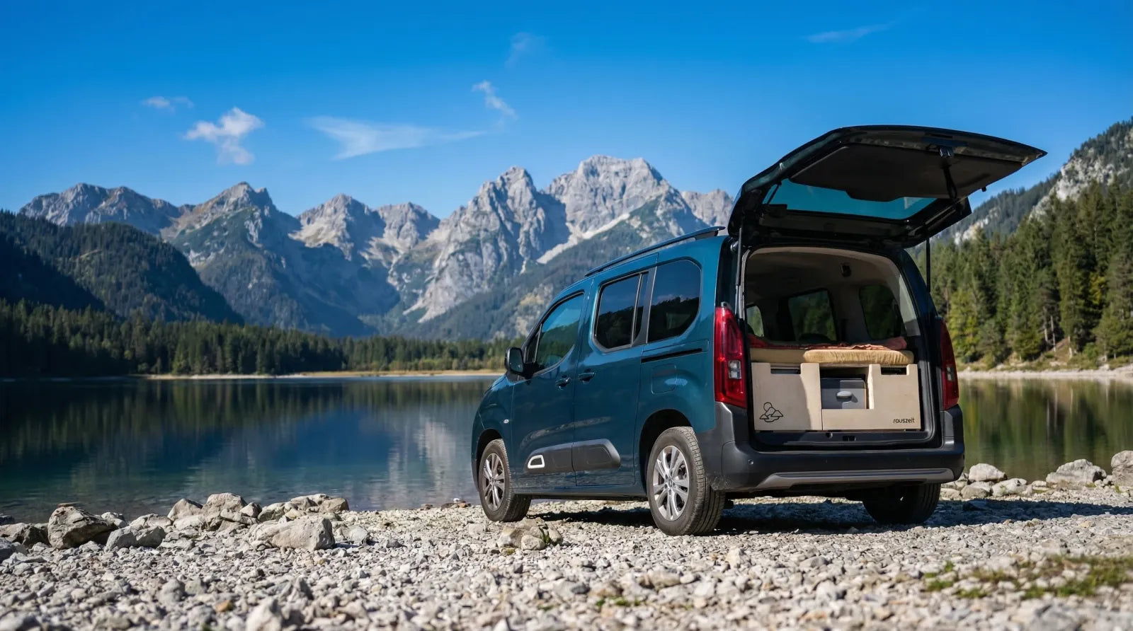 Citroën Berlingo mit Campingbox am steinigen Seeufer vor Bergkulisse bei blauem Himmel