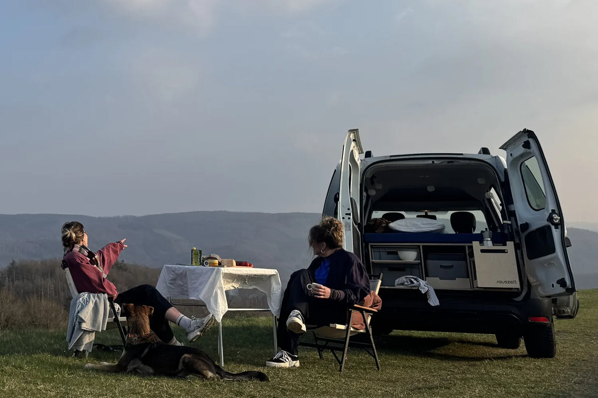Dacia Dokker mit offenen Hecktüren und Rauszeit Campingbox auf einer Wiese in der Eifel, davor zwei Frauen mit Hund am Campingtisch