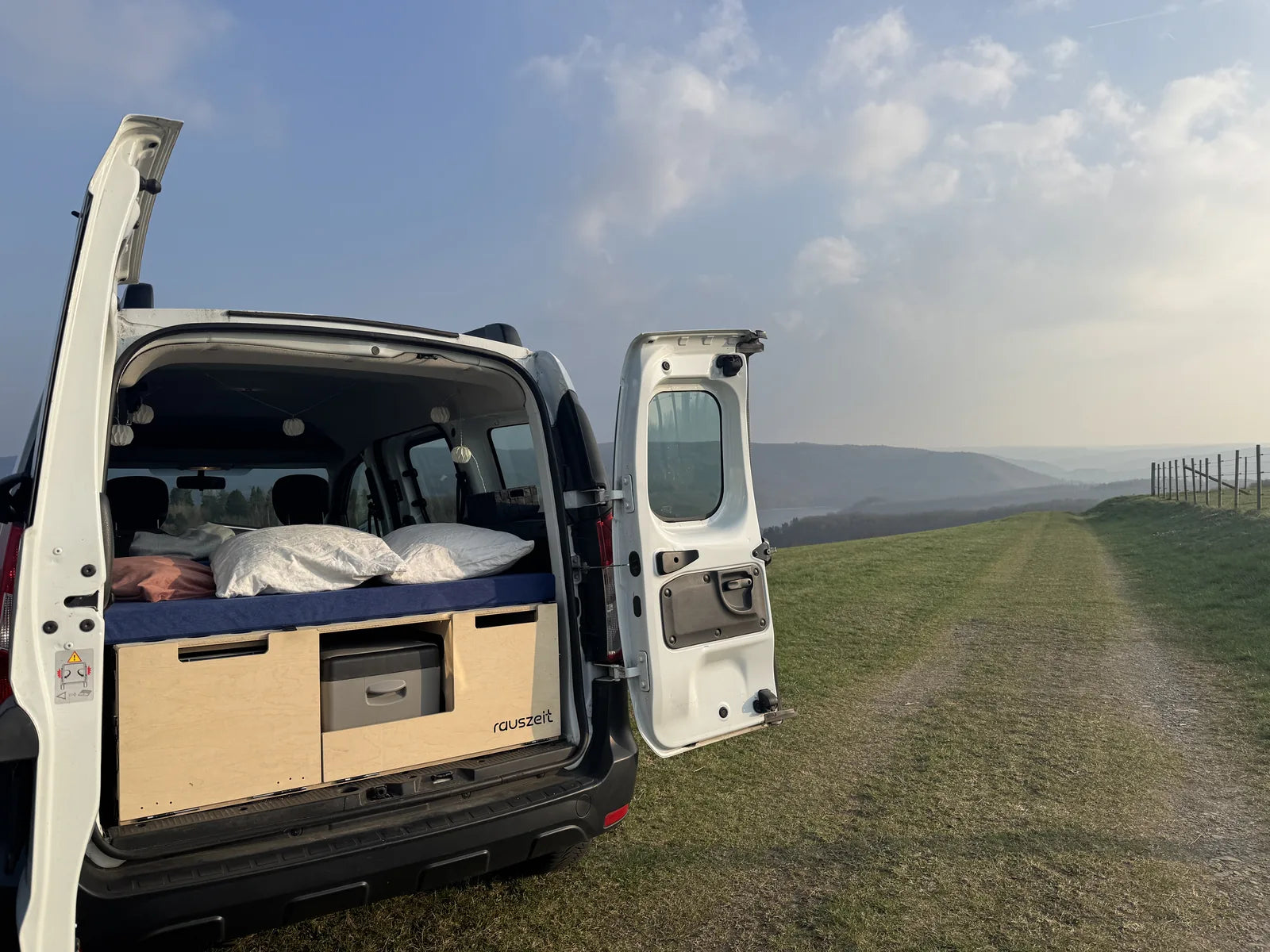 Offenes Heck eines weißen Dacia Dokker mit Rauszeit Campingbox und gemachtem Bett, Blick über die Eifel