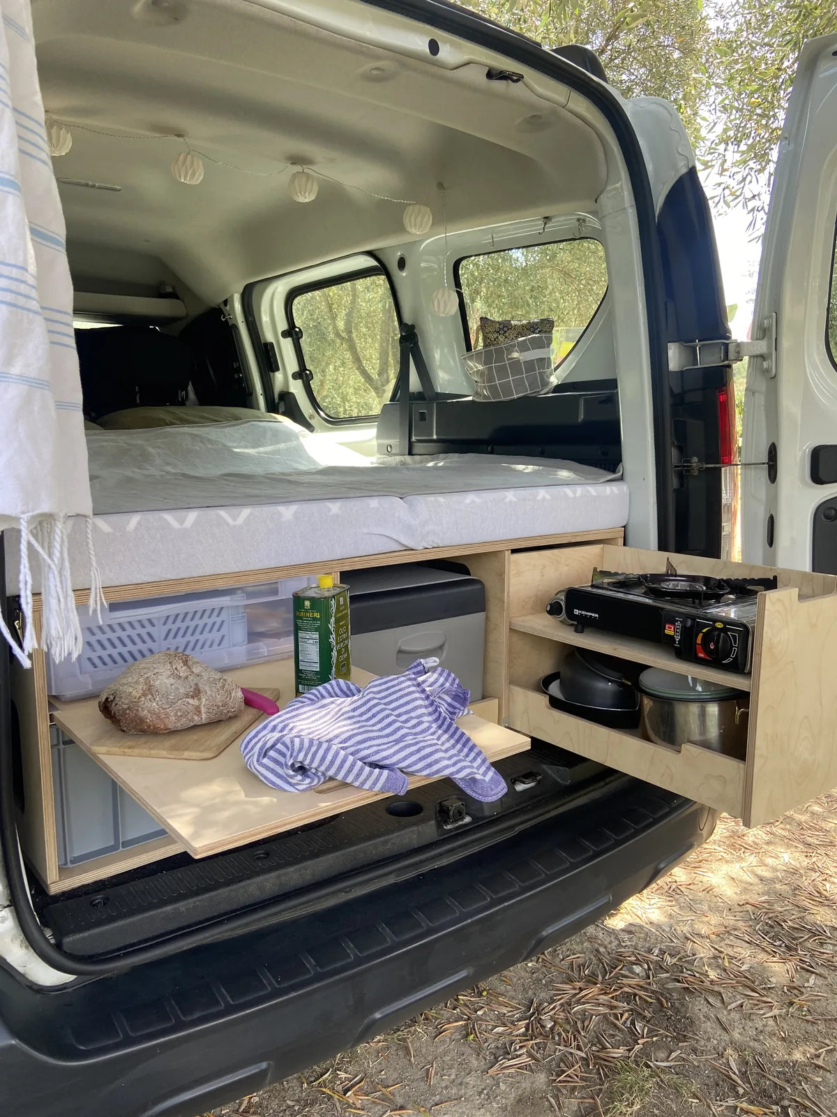 Offenes Heck eines Dacia Dokker: Rauszeit Campingbox mit ausgezogenem Kochmodul, Gaskocher, Töpfen und Schneidebrett, darüber das Bett
