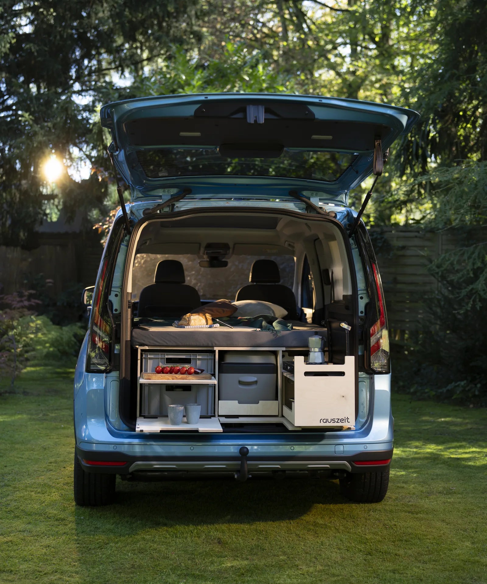 Hellblauer Ford Tourneo Connect von hinten mit offener Heckklappe, darin die Rauszeit Campingbox mit Kochmodul, Euroboxen und Kühlbox