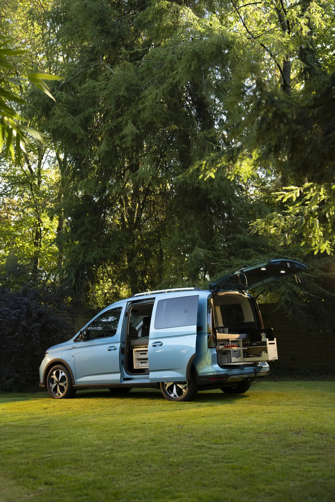 Ford Tourneo Connect mit offener Schiebetür und Heckklappe unter hohen Bäumen, Campingbox eingebaut