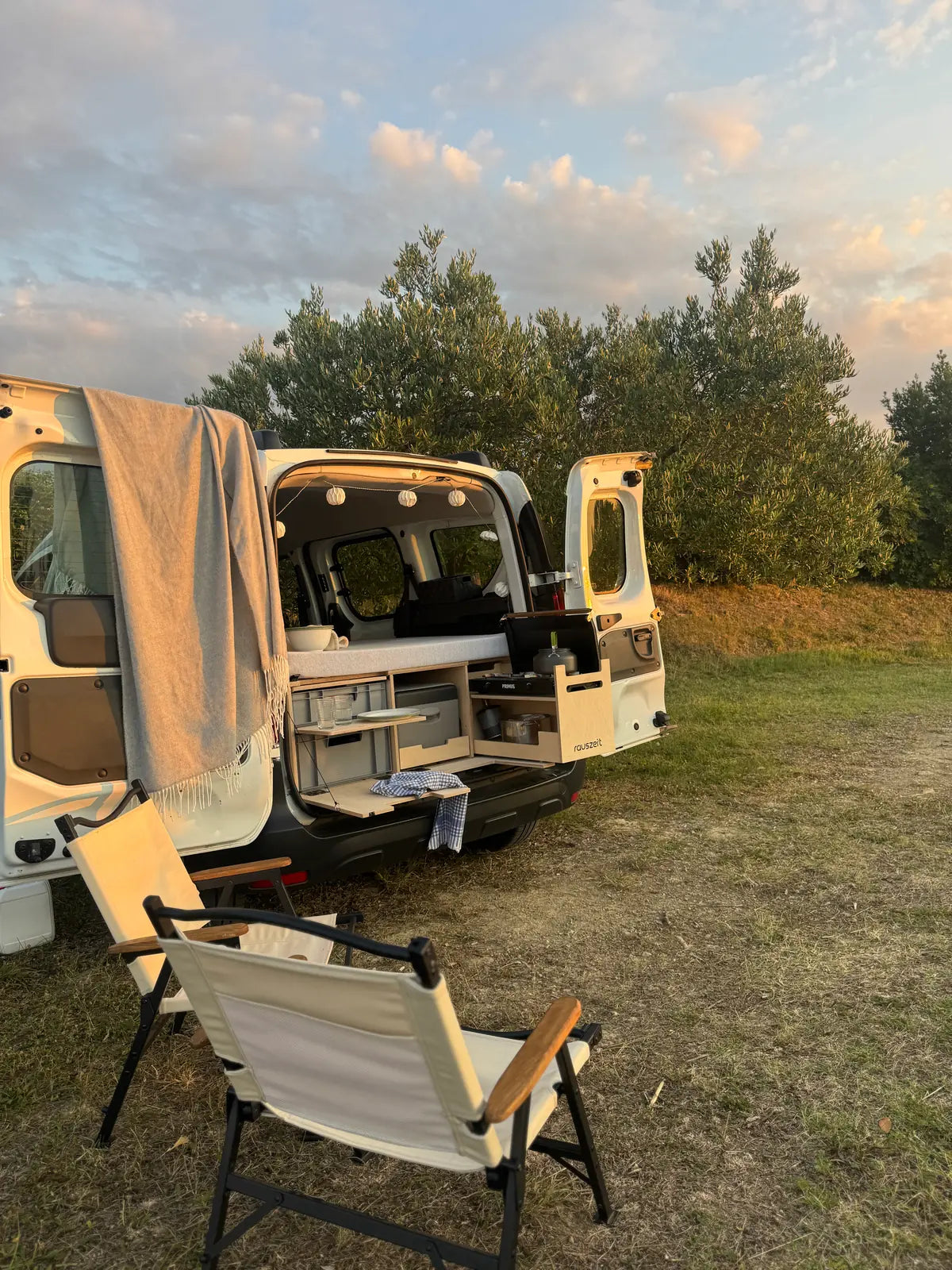 Campingbox mit offenen Heckklappen, sichtbarem Stauraum und Campingstuhl auf einer Wiese bei Abendstimmung