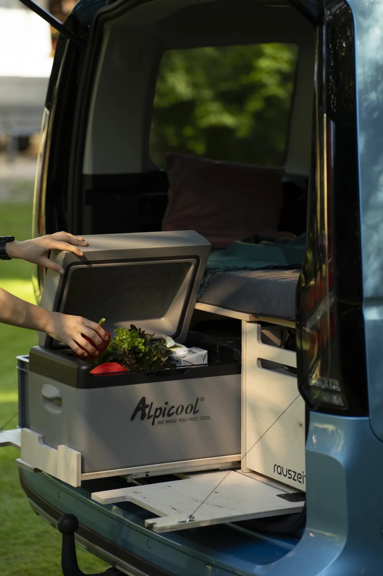 Kühlbox auf dem ausgezogenen Auszug der Campingbox am offenen Heck, eine Hand nimmt Gemüse heraus
