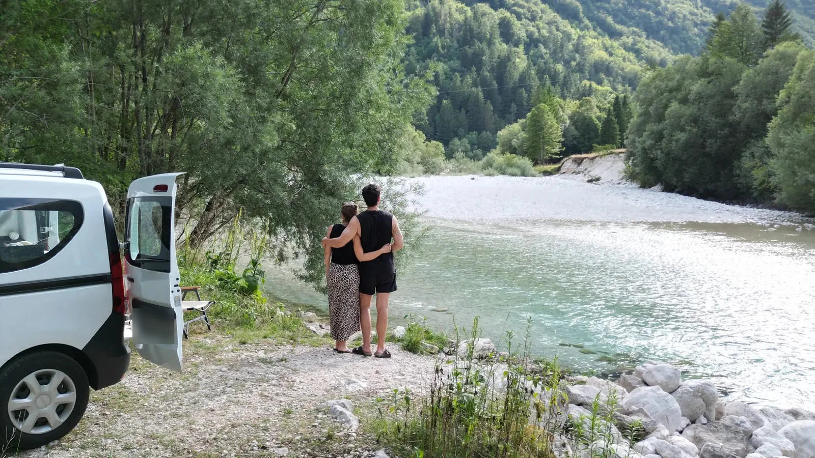 Paar am türkisfarbenen Soča-Fluss neben weißem Dacia Dokker vor grüner Bergkulisse