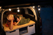 Paar liegt abends auf dem Schiebebett der Campingbox mit Lichterkette