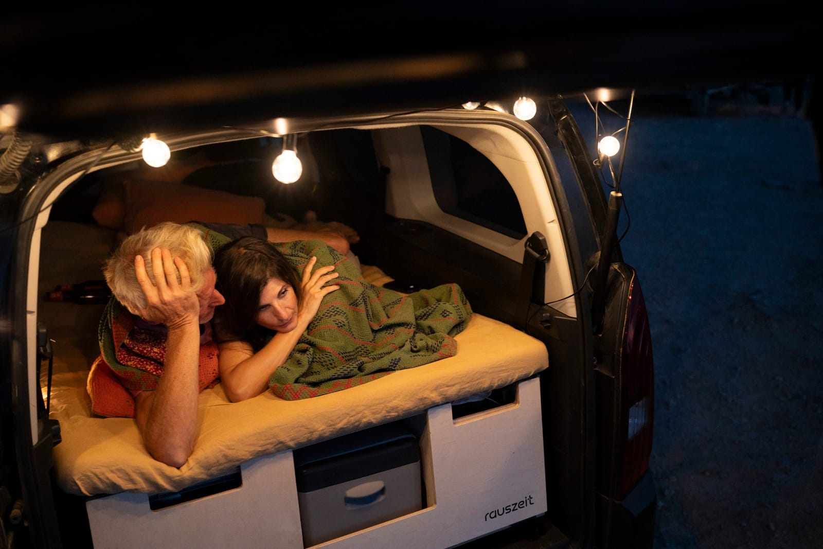 Paar liegt abends auf dem Schiebebett der Campingbox mit Lichterkette